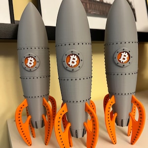 May include: Three grey rocket models with orange fins. Each rocket features a Bitcoin logo in a circular emblem. The rockets are displayed on a white surface, with a framed image in the background.