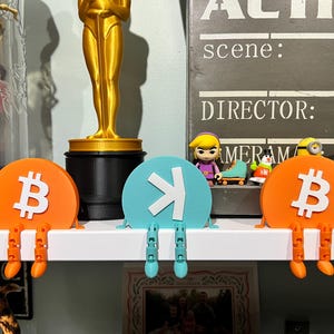 Puede incluir: Tres soportes de estante impresos en 3D, cada uno con un diseño diferente. El primero es naranja con un símbolo de Bitcoin blanco, el segundo es azul con una flecha blanca que apunta hacia la derecha, y el tercero es naranja con un símbolo de Bitcoin blanco. Los soportes de estante están en un estante blanco.