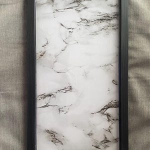May include: A rectangular serving tray featuring a white surface with a grey marble pattern. The tray is enclosed by a solid black frame and includes two brushed metal handles attached to the top and bottom edges for carrying.
