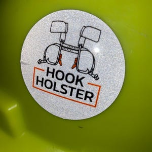 Op de afbeelding: Een witte reflecterende sticker met een zwart-witte illustratie van een gereedschapsgordel met de tekst "HOOK HOLSTER" in een rood rechthoek.