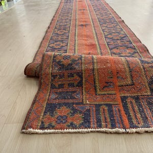 Peut inclure: Un tapis de couloir rectangulaire enroulé, d'aspect vintage et vieilli. Le tapis présente une bande centrale orange flanquée de bordures à motifs bleu foncé et orange. Les bords du tapis ont une frange crème.