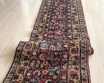 Puede incluir: Una alfombra de pasillo rectangular enrollada con un patrón floral en tonos de rojo, azul, dorado y negro. La alfombra está colocada sobre un suelo de madera clara, mostrando su intrincado diseño y su rica paleta de colores.