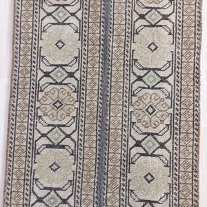 May include: A rectangular runner rug featuring a repeating geometric pattern in shades of beige, tan, and dark charcoal gray. The design consists of vertical bands with octagonal motifs and intricate linear borders, creating a symmetrical layout.