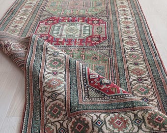 May include: A traditional rug with a green, red and cream colour scheme. The rug has a geometric pattern with intricate details. The rug is folded in half, revealing the underside.
