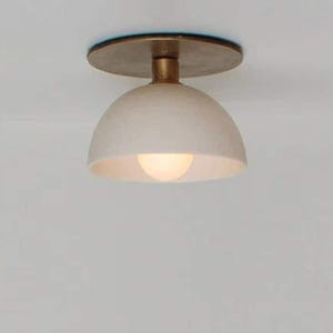 May include: A modern ceiling light fixture with a cream-colored, dome-shaped shade and a brass-colored stem and mounting plate. The light bulb is visible inside the shade, emitting a warm glow. The fixture is designed for indoor use.