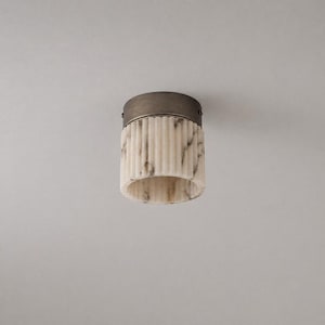 Op de afbeelding: Een cilindervormige plafondlamp met een geribbelde witte marmeren kap met grijze aders. De bovenkant van het armatuur is afgedekt met een ronde, donker metalen bronzen montageplaat die is vastgezet met kleine schroeven aan de zijkant.