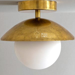 May include: A brass-coloured ceiling light fitting with a rounded shade and a white globe-shaped light. The fitting has the text "Brass Light" and a lightbulb icon. The light is designed to be mounted on a ceiling.