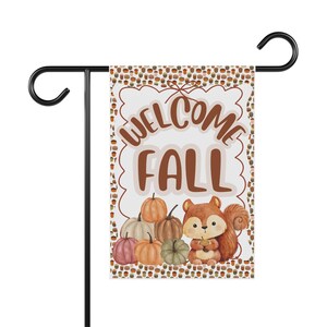May include: A rectangular garden flag hanging on a black metal stand. The flag features a white center with the brown text "WELCOME FALL" above a cluster of pumpkins and a squirrel holding an acorn. The border is patterned with small acorns.