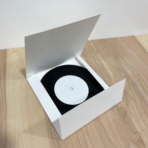 May include: White cardboard record sleeve with a black vinyl record inside. The record has a white label with a small hole in the center.