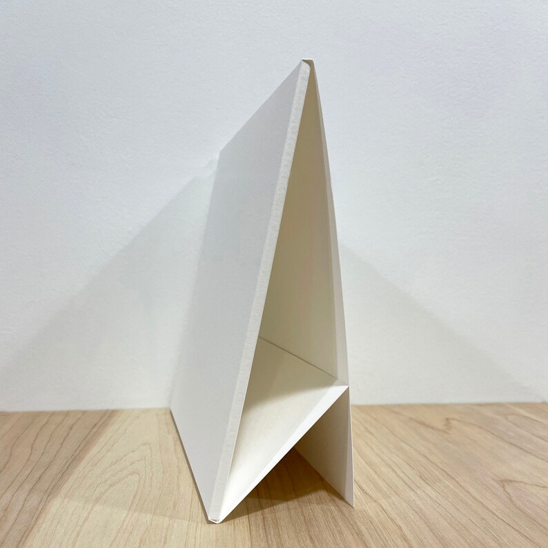 May include: A white cardboard easel stand with a triangular shape. The stand is made of thick cardboard and has a sturdy construction.