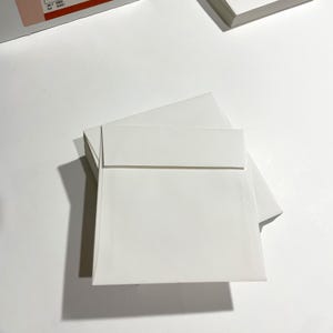 May include: A stack of white square envelopes with a flap closure. The envelopes are arranged on a white surface, with a shadow cast to the left. The image is well-lit, highlighting the clean, minimalist design of the stationery.