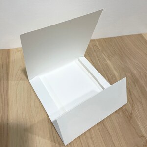 May include: A white card with a flap that folds open to reveal a square opening. The card is made of thick paper and has a smooth finish.