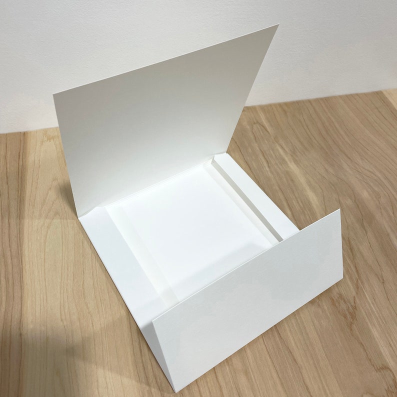 May include: A white card with a flap that folds open to reveal a square opening. The card is made of thick paper and has a smooth finish.