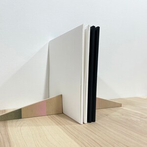 May include: A white book with a black spine is standing upright on a wooden shelf. The book is propped up by a wooden wedge with a pink and green stripe.