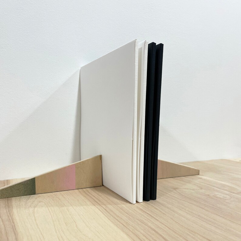 May include: A white book with a black spine is standing upright on a wooden shelf. The book is propped up by a wooden wedge with a pink and green stripe.