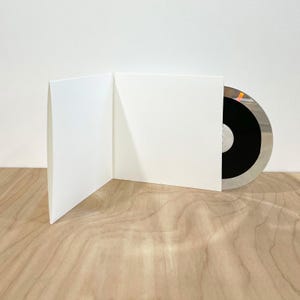 May include: A white cardboard CD gatefold case standing open, revealing a compact disc with a black center and a reflective silver outer ring. The disc is partially inserted into the right side of the plain, unprinted white paper sleeve.