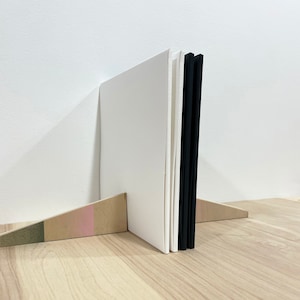 May include: A white book with a black spine is propped up on a wooden stand with a pink and green stripe. The book is standing on a light wood surface.