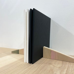 May include: Three books standing upright on a wooden shelf. The books are bound in black and white paper. The books are propped up by a wooden wedge.