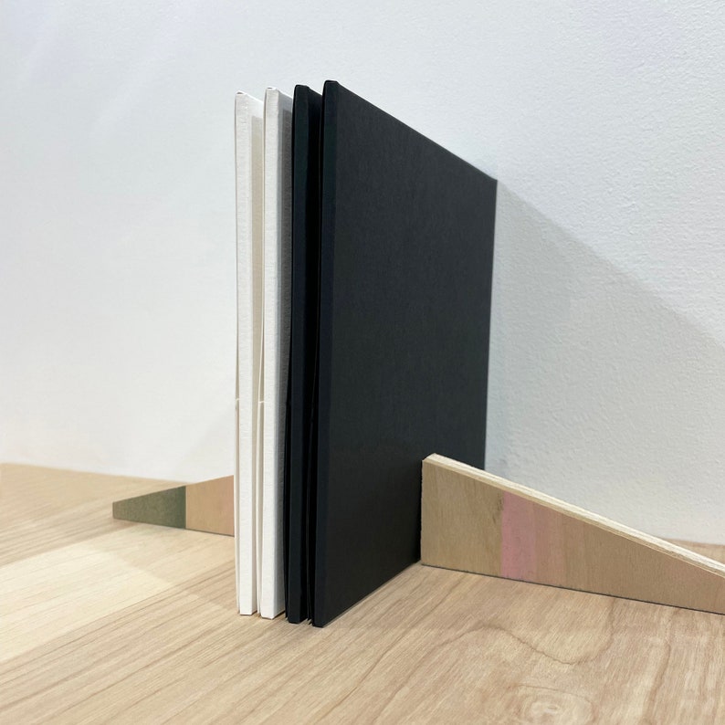 May include: Three books standing upright on a wooden shelf. The books are bound in black and white paper. The books are propped up by a wooden wedge.