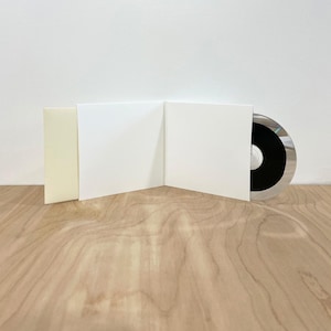 May include: A white cardboard CD gatefold case with a matte finish, featuring a blank interior and a single disc partially inserted into the right panel. The disc has a black center label with a reflective silver outer ring and clear plastic edge.