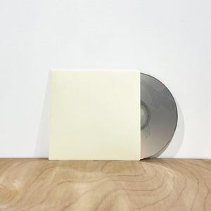 May include: A silver CD partially inserted into a square, off-white paper sleeve. The CD has a perforated design. The sleeve is positioned on a light-colored wooden surface against a white wall.