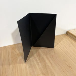 May include: A black, folded paper object with a geometric design. The paper is folded to create a triangular shape on one side, with the other side open. The object is on a light wood surface.