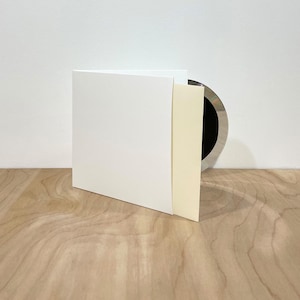 May include: A square white cardstock folder with a cream-colored insert partially pulled out, revealing a circular metallic silver disc with a black center partially visible behind the paper elements, creating a layered and minimalist composition.