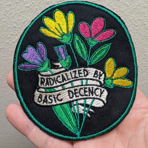 May include: A circular embroidered patch with a black background and a green border. It features a bouquet of yellow, pink, and purple flowers with green stems. A white ribbon banner across the center displays the text RADICALIZED BY BASIC DECENCY.