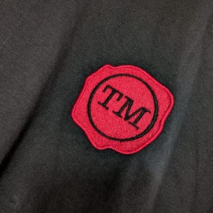 May include: A black fabric garment featuring a circular red embroidered patch with a scalloped edge. The patch contains a black circular border and the black embroidered letters TM in the center, resembling a wax seal design on the dark material.