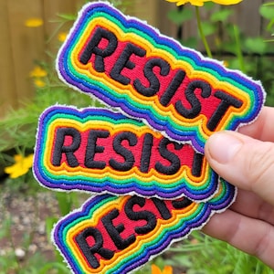 Może przedstawiać: Haftowane naszywki ze słowem "RESIST" czarnymi literami. Naszywki mają tęczową obwódkę i czerwone tło. Naszywki są ułożone jedna na drugiej.