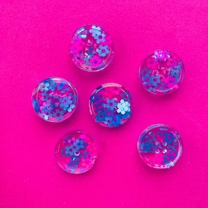 May include: Six clear resin buttons with a pink and blue glitter embedded inside. The buttons have a flower design and are arranged on a bright pink background.