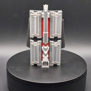 May include: A silver and red prop device, possibly a wrist-mounted gadget, with rectangular panels and a central vertical component. The device is mounted on a black rotating platform, suggesting a display or presentation setting. The design appears to be futuristic or inspired by science fiction.