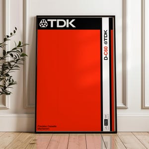 May include: A framed poster featuring a vibrant red background with a black and white stripe on the right side. The top section is black with the white text "TDK" and a logo. The text "D-C60 TDK" is on the white stripe. The bottom text reads "Precision Cassette Mechanism".