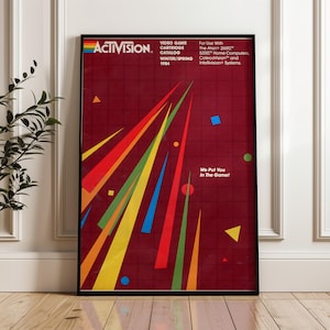 Puede incluir: Póster enmarcado del catálogo de cartuchos de videojuegos Activision de invierno/primavera de 1984. El póster presenta un fondo burdeos con formas y líneas geométricas coloridas. El texto incluye "We Put You In The Game!" e información de compatibilidad.