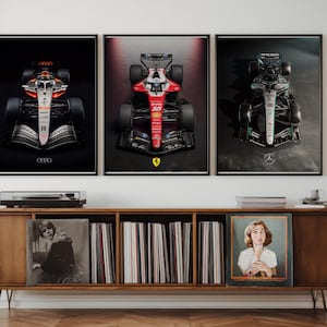 May include: Three framed posters of Formula 1 race cars. The posters show close-up views of the cars against a dark backdrop. The cars are in various colours, including red, silver, and black. The posters are displayed above a wooden cabinet with a record player and vinyl records.