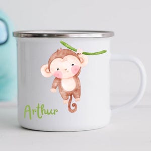 Puede incluir: Taza de esmalte blanco con un mono de dibujos animados colgado de una vid. La taza tiene un borde y un asa plateados. El texto "Arthur" está impreso en verde debajo del mono.