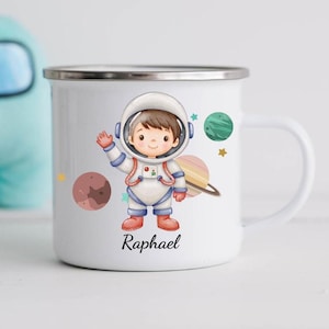 Peut inclure: Mug en émail blanc avec une illustration de dessin animé d'un garçon astronaute aux cheveux bruns et une combinaison spatiale rouge et bleue. La tasse a une planète bleue et verte, une planète rouge et des étoiles. Le texte "Raphael" est imprimé sur la tasse.