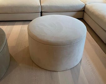 May include: A large, round, off-white ottoman with a smooth, upholstered surface. The ottoman is sitting on a light wood floor.