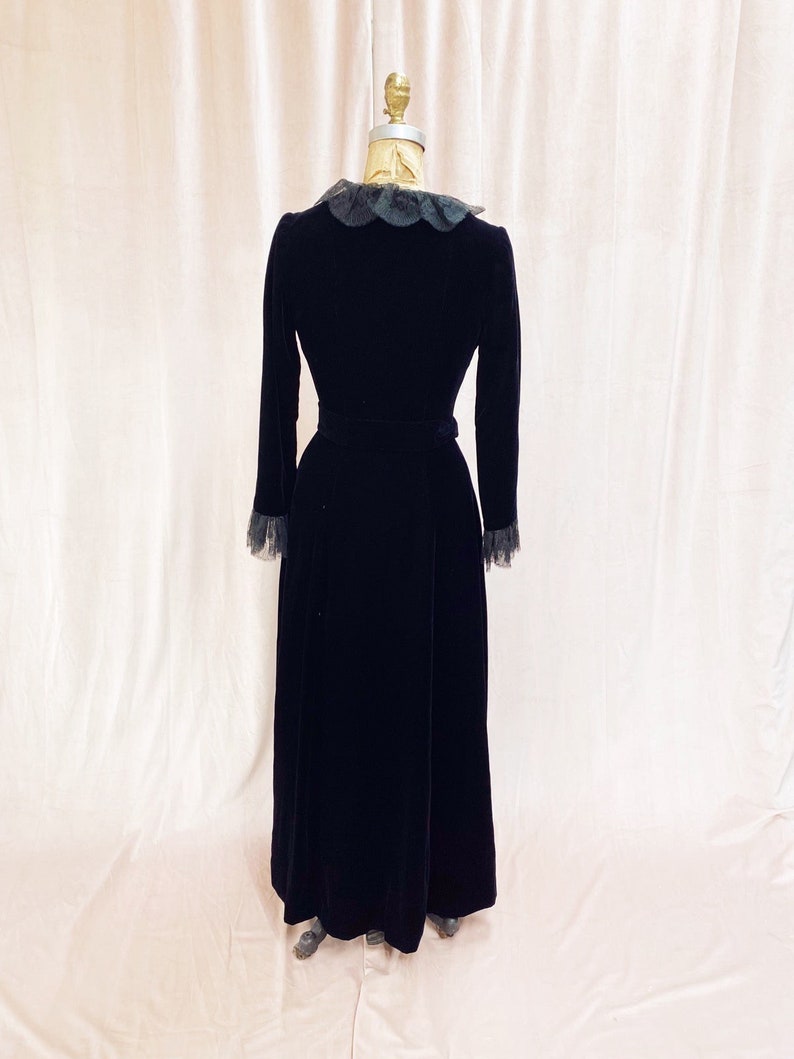 Pode incluir: Um vestido maxi de veludo preto com colarinho e punhos de renda. O vestido tem um corpete justo e uma saia fluida.