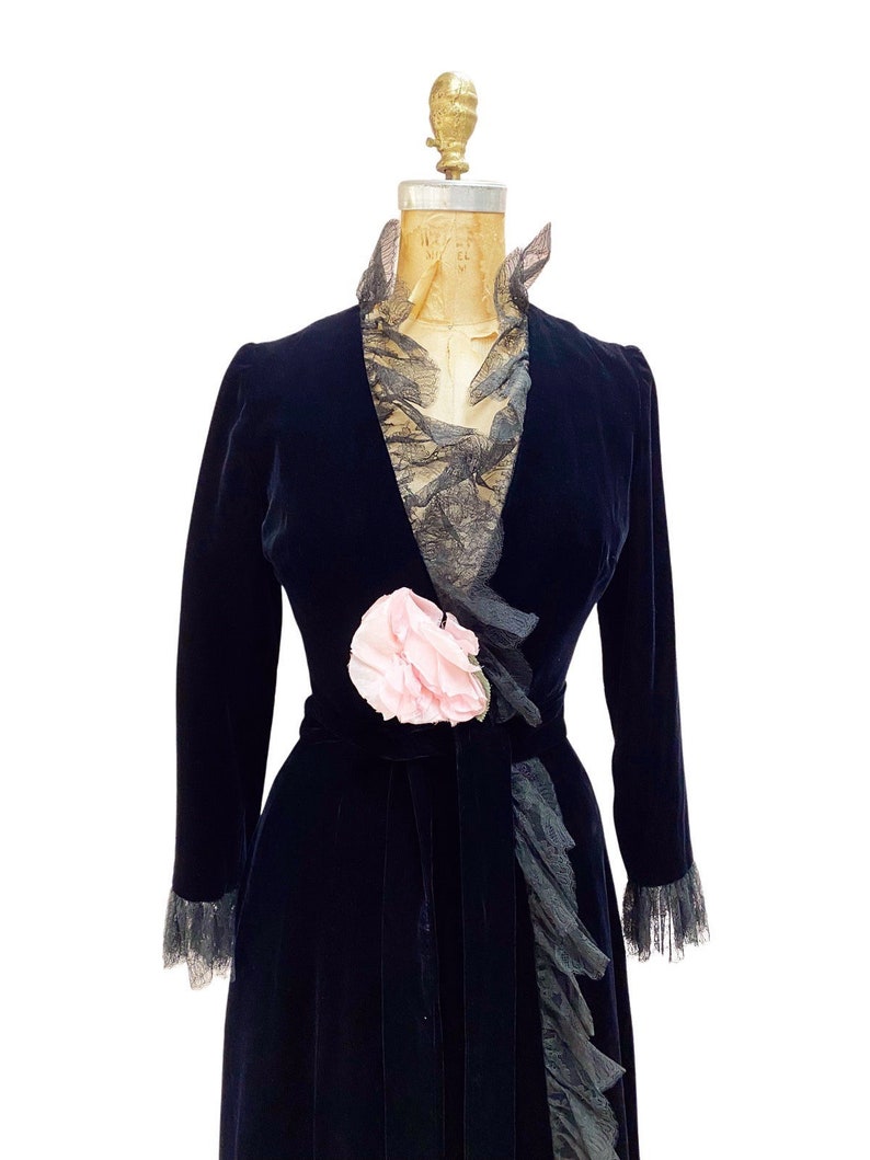 Pode incluir: Uma longa robe de veludo preto com gola de renda e um detalhe de flor rosa na cintura.