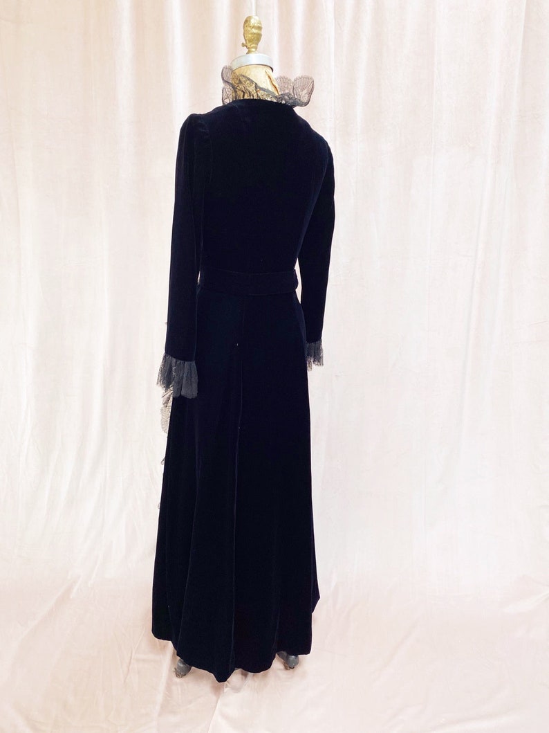Pode incluir: Um vestido longo de veludo preto com colarinho e punhos de renda. O vestido tem um corpete justo e uma saia fluida.