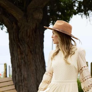 Könnte beinhalten: Ein hellbrauner Fedora-Hut mit geflochtenem Band und breiter Krempe. Der Hut wird von einer Person getragen, die ein cremefarbenes Kleid mit Spitzendetails an den Ärmeln trägt. Der Hintergrund umfasst einen großen Baum und eine Holzbank.