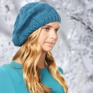 Può includere: Un berretto di lana blu turchese con un motivo a treccia. Il berretto è indossato da una persona con lunghi capelli biondi.