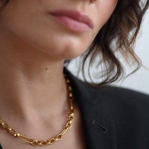 Könnte beinhalten: Eine goldfarbene Kette mit einem Kettendesign. Die Halskette wird vor einem dunklen Blazer getragen. Das Bild ist eine Nahaufnahme, die sich auf die Halskette sowie den Hals und das Gesicht der Person konzentriert.