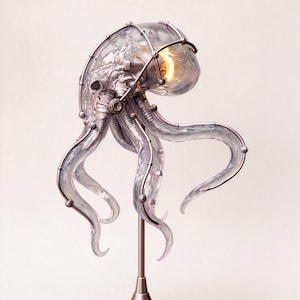 May include: A sculptural table lamp shaped like a jellyfish with a clear, translucent dome and flowing tentacles. The structure is encased in a metallic, silver-toned framework with visible mechanical joints and a warm, glowing light source.