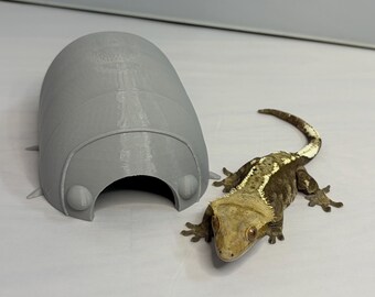 Peut inclure: Un abri pour reptile gris imprimé en 3D, de forme arrondie avec de petits détails en saillie. Un gecko à crête avec un motif marron et beige est positionné à côté de l'abri, présentant un accessoire pour animaux de compagnie.