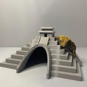 May include: A gray, 3D-printed reptile habitat shaped like a stepped pyramid with an arched entrance and a small temple-like structure on top. A gecko with yellow and brown skin is perched on the side.