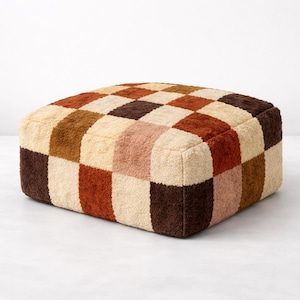 Peut inclure: Un pouf carré et touffeté avec un motif en damier. Les carrés sont dans des tons crème, marron, rouille et rose. Le pouf est un meuble bas, au niveau du sol.