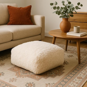 Op de afbeelding: Een crèmekleurige, vierkante poef met een zacht, getextureerd oppervlak staat op een patroonkleed. Een roestkleurig kussen ligt op een beige bank. Een houten salontafel heeft een vaas en een kleine beker.