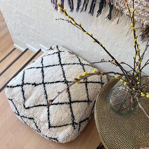 Puede incluir: Un cojín de suelo blanco y negro con un diseño geométrico. El cojín está hecho de un material suave y esponjoso y está sentado en un suelo de madera. Un jarrón de flores amarillas está en el fondo.